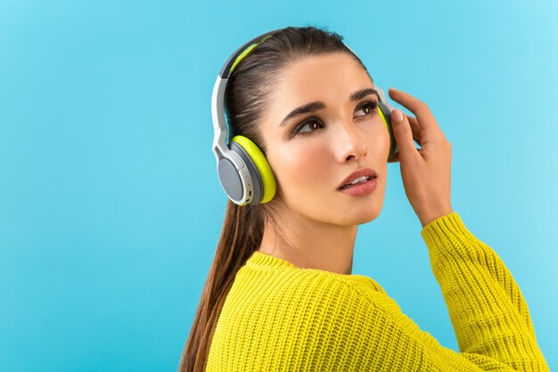 Attractive stylish young woman listening to music in wireless headphones happy wearing yellow knitted sweater colorful style fashion posing isolated on blue wall