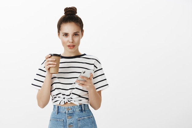 スマートフォンを使用して、カフェからコーヒーを飲みながら魅力的なスタイリッシュな若い女性