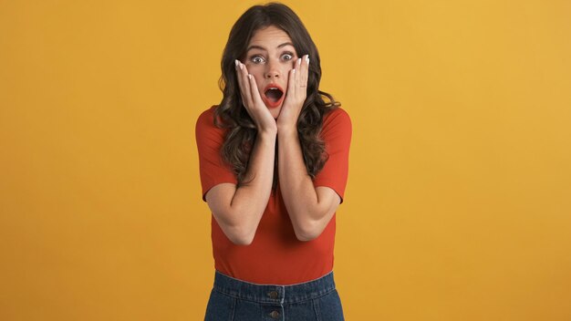 Attractive surprised brunette girl in red top amazedly looking in camera over colorful background Wow expression