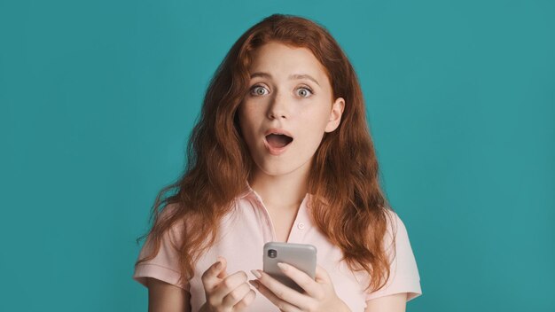 Attractive surprised redhead girl with smartphone amazedly looking in camera with open mouth over colorful background Wow expression