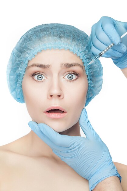 Attractive surprised and scared woman at plastic surgery with syringe in her face on white background