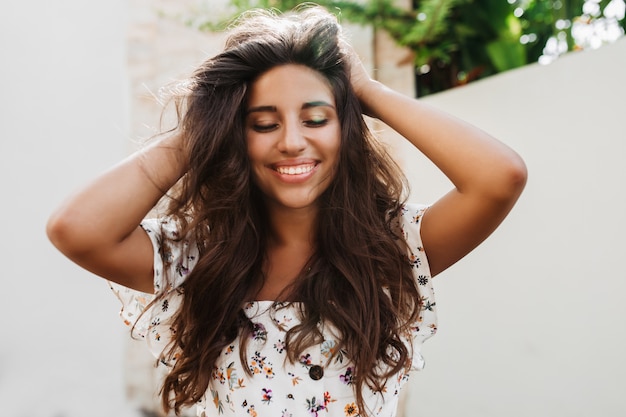 Attractive tanned woman in white blouse with charming smile posing on white wall with green plants