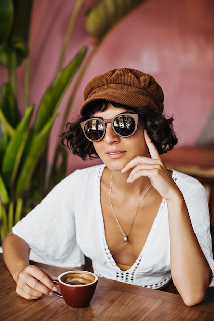 Attraente donna abbronzata in maglietta di cotone bianco e berretto marrone sorridente e tenendo la tazza di caffè con latte