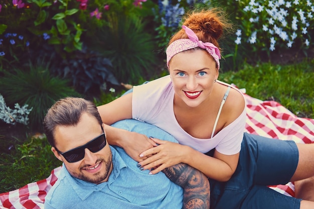 Free photo attractive tattooed, bearded male and a redhead female lying on a blanket on a lawn in a park. picnic time.