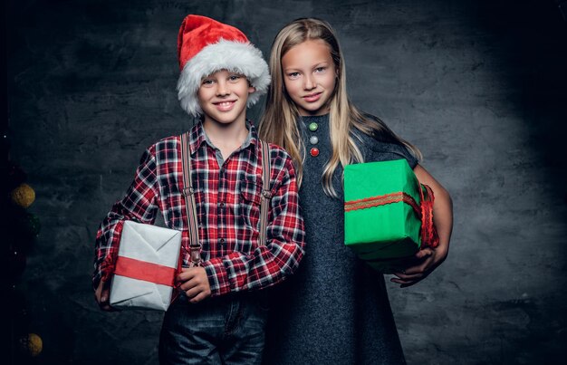 Attractive teenage boy on Santa's hat and cute blonde girl holds Christmas gift boxes.