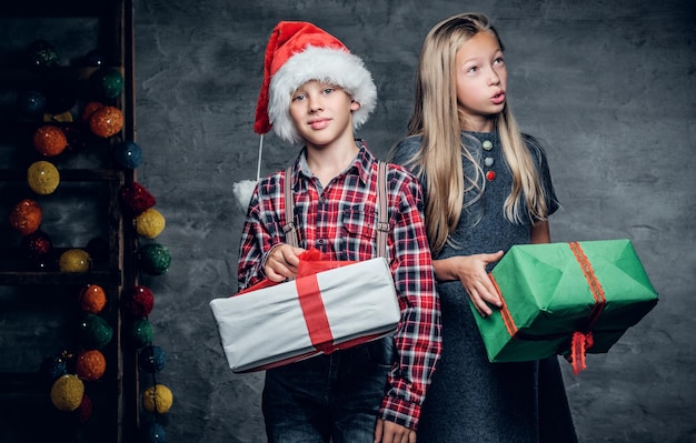 Attractive teenage boy on Santa's hat and cute blonde girl holds Christmas gift boxes.