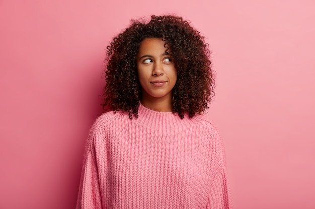 아프로 머리를 가진 매력적인 십대 소녀는 오른쪽 상단 모서리에 잠겨있는 것처럼 보이고 분홍색 스웨터를 입은 사려 깊은 표정을 가지고 실내 포즈를 취하고 무언가에 대한 의구심을 가지고 있습니다.
