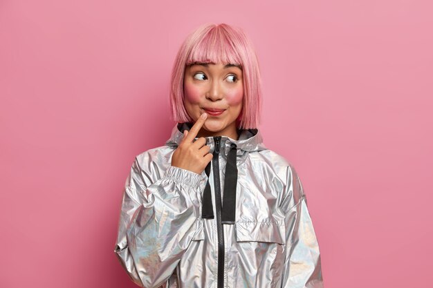 attractive teenage girl with trendy pink hairstyle, keeps index finger near lips, looks curiosly aside dressed in fashionable silver coat 