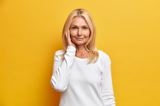 Attractive tender middle aged woman with blonde hair has healthy and wrinkled skin wears minimal makeup dressed in casual white jumper poses indoor 