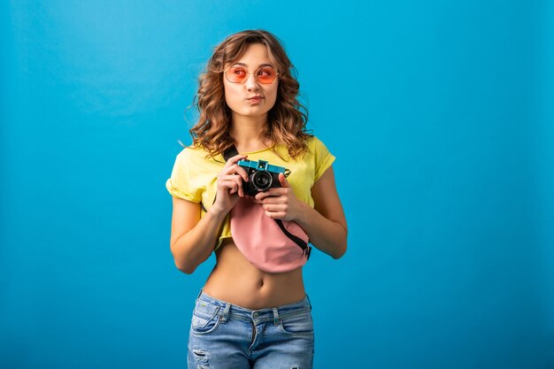 Привлекательная женщина мышления позирует со старинной фотокамерой, фотографируя, одетая в красочный летний хипстерский костюм, изолированные на синем фоне студии