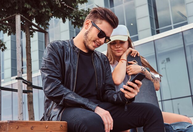 Attractive trendy dressed young couple have fun together while sitting together on bench near skyscraper.