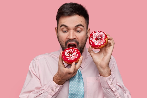 Attractive unshaven businessman being hungry after hard work, bites delicious red doughnut, enjoys nice taste, wears formalwear