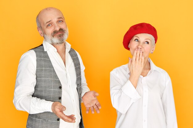 Attractive unshaven man with bald head having frustrated mournful expression, making helpless gesture, his beautiful stylish mature wife looking up in surprise, covering mouth with hand, gasping