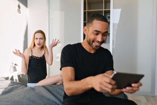 Attractive upset funny young woman on bed is angry at excited happy guy playing with tablet on bed at home in modern apartment. Relationship, offended girl, having fun, incomprehension