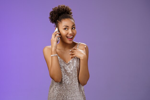 Attractive wealthy talkative pleased african american girl discuss silly things calling friend smartphone speaking phone gesturing pointing herself bragging during phonecall standing blue background.