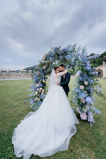 Attractive wedding couple in love is standing outdoors near the beautiful archway made of blue flowers