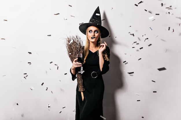 Attractive witch holding wineglass with blood. Indoor photo of blonde lady in wizard costume posing under confetti in halloween.