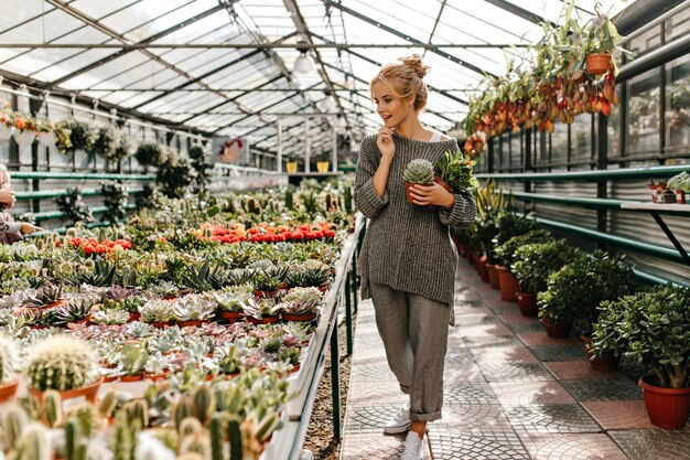 헐렁한 세련된 바지와 스웨터를 입은 매력적인 여성은 집에 식물을 선택하고 즙을 유지합니다.