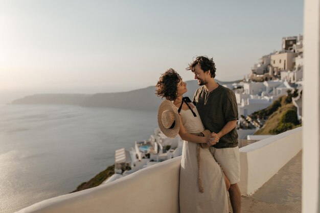 Attractive woman in beige dress boater and brunette man hold hands and look at each other with love Couple poses in beautiful romantic place with sea view