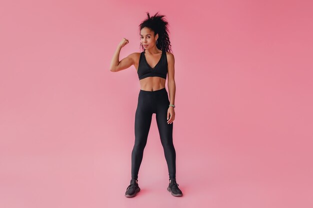 Attractive woman in black leggings and top fitness outfit on pink isolated wall