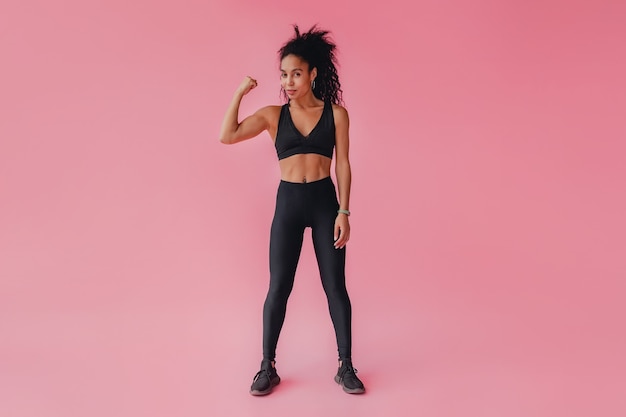 Attractive woman in black leggings and top fitness outfit on pink isolated wall