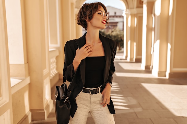 Attractive woman in black top, jacket and white trousers with belt posing outside. Wavy-haired woman with handbag and glasses looks away in city.