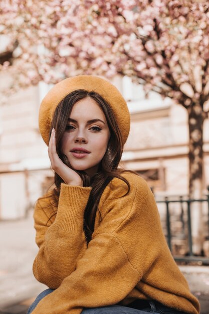 Attractive woman in bright beret and sweater looks into camera against background of building. Pretty young brunette lady in good mood posing in city against sakura