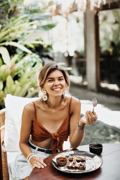 Attractive woman in brown bra smiles widely and eats tasty waffle with ice cream and chocolate sauce