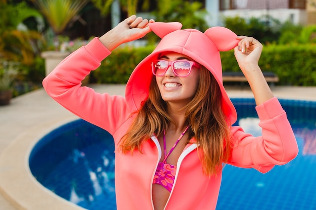 Привлекательная женщина в красочной розовой толстовке с капюшоном в солнцезащитных очках на летних каникулах, улыбаясь эмоциональным выражением лица, весело, спортивный стиль моды