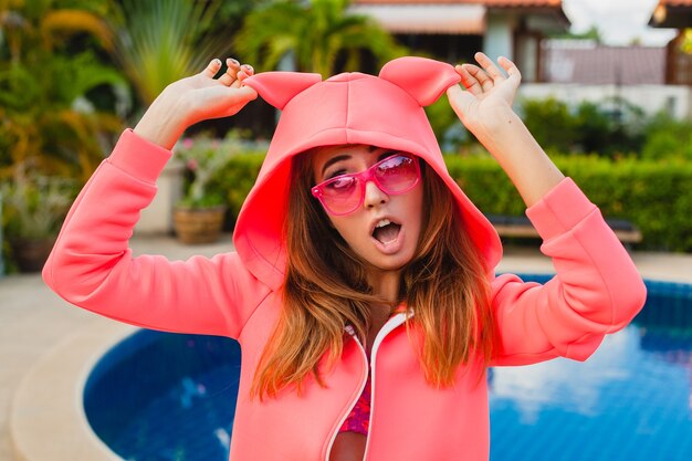 Привлекательная женщина в красочной розовой толстовке с капюшоном в солнцезащитных очках на летних каникулах, улыбаясь эмоциональным выражением лица, весело, спортивный стиль моды
