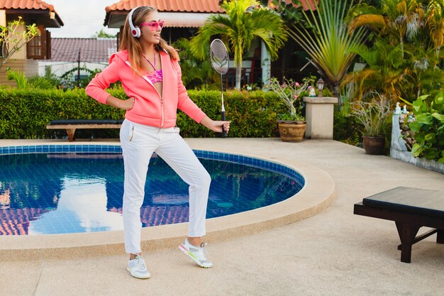 Attractive woman doing sports at pool in colorful pink hoodie wearing sunglasses listening to music in headphones on summer vacation, play tennis, sport style