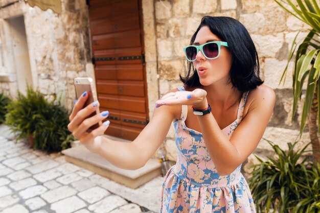 Привлекательная женщина в платье, путешествуя на отдыхе в старом центре города Италии, делает смешное селфи на телефоне, отправляя поцелуй