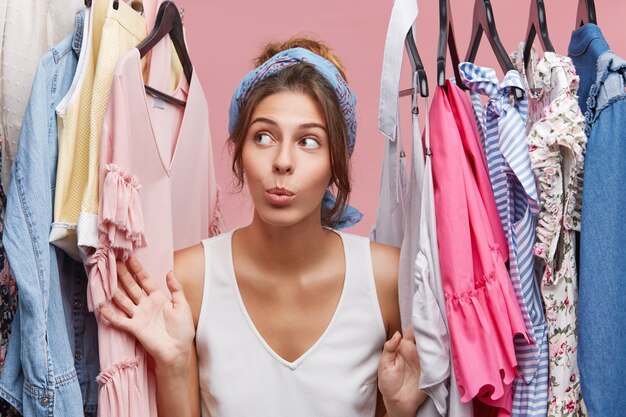 Attractive woman dressed casually, looking with doubts aside while standing near hangers with clothes, thinking what to dress on business meeting with companions. Woman of fashion having many clothes