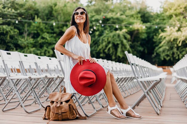 白いドレス、赤い帽子、椅子だけで夏の野外劇場に座っているサングラス、春のストリートスタイルのファッショントレンド、アクセサリー、バックパックと一緒に旅行に身を包んだ魅力的な女性