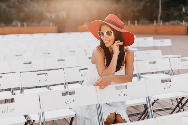 Привлекательная женщина, одетая в белое платье, красную шляпу, солнцезащитные очки, сидя в летнем театре под открытым небом на стуле в одиночестве, весенняя уличная мода, социальное дистанцирование