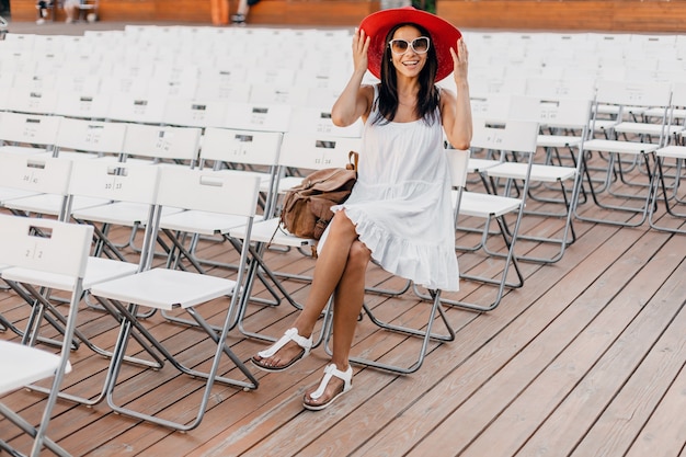 Attractive woman dressed in white dress, red hat, sunglasses sitting in summer open air theatre on chair alone, spring street style fashion trend, social distancing