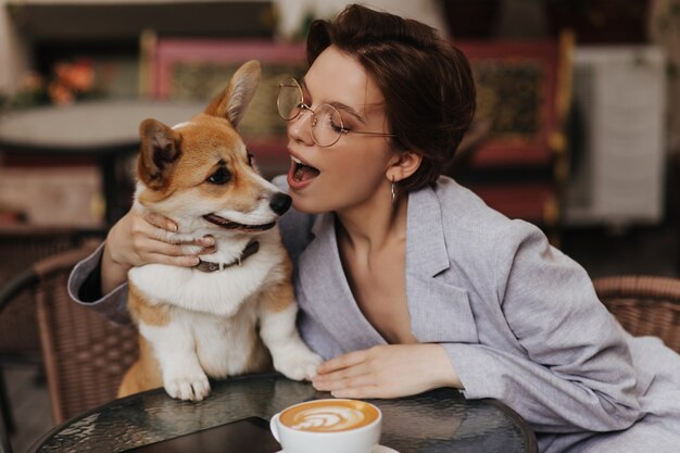 Привлекательная женщина в очках играет со своей собакой во время отдыха в уличном кафе. Очаровательная дама в сером пиджаке целует корги