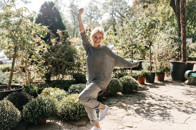 Attractive woman in gray jeans pants is having fun in park Girl blonde in sweater posing with smile