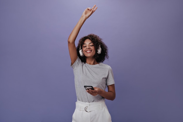 Привлекательная женщина в серой футболке наслаждается музыкой и держит смартфон Брюнетка в белых шортах танцует на фиолетовом фоне