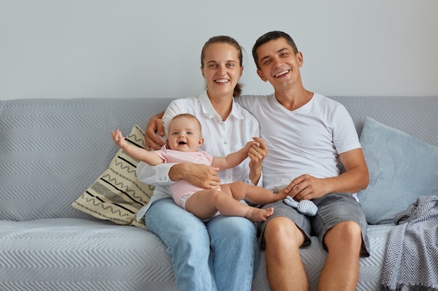 Attractive woman and handsome female sitting on sofa with infant daughter, looking smiling at camera, being happy together, family at home, indoor shot.