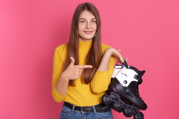 Free photo attractive woman holding roller skating in hands and pointing at it with her index finger, , posing against rosy wall.