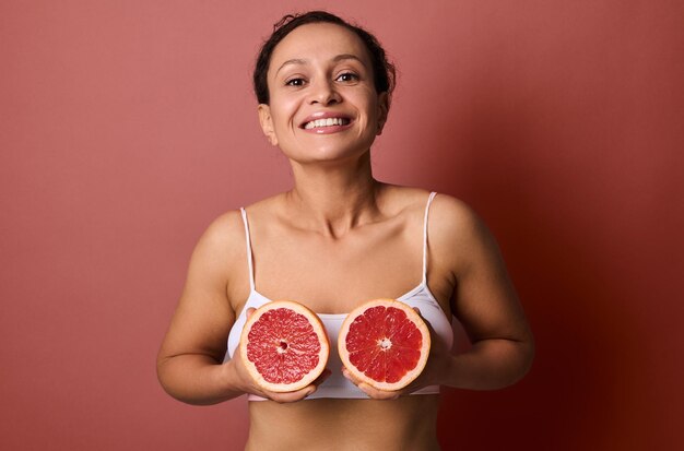 Привлекательная женщина в белом нижнем белье держит половинки грейпфрутов на уровне груди, сладко улыбаясь зубастой улыбкой, позируя на коралловом фоне с местом для текста и рекламы. уход за телом и кожей