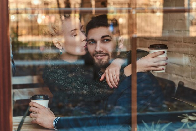 Attractive woman is kissing her man while sitting at cafe with cup of coffee.