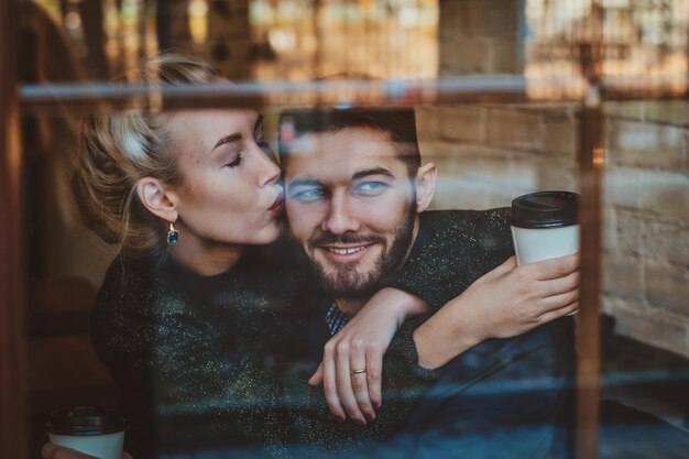 매력적인 여자가 커피 한잔과 함께 카페에 앉아있는 동안 그녀의 남자에게 키스하고 있습니다.