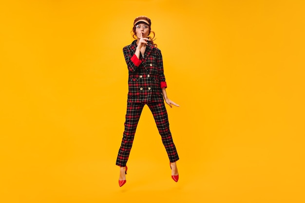 Attractive woman jumps on orange background and asks to keep secret Fashionable girl in brown trendy cap and plaid suit posing