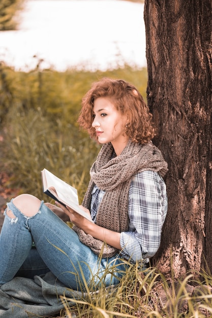 Attractive woman leaning on tree and holding book in park