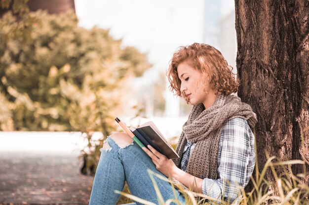 Attractive woman leaning on tree and reading book in sunny park