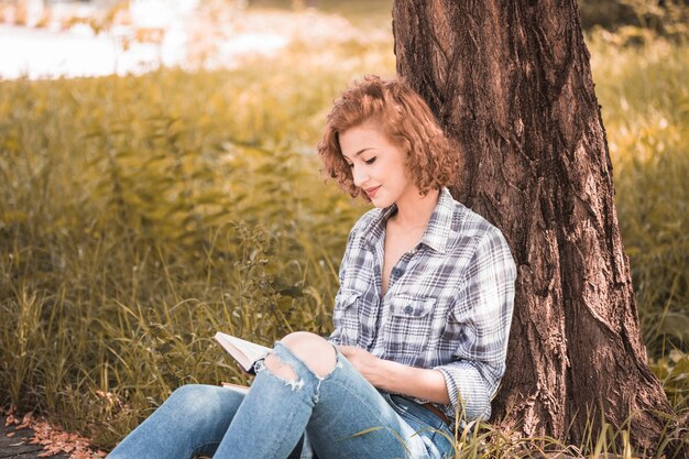 매력적인 여자가 나무에 기대어 공공 정원에서 책을 낭송