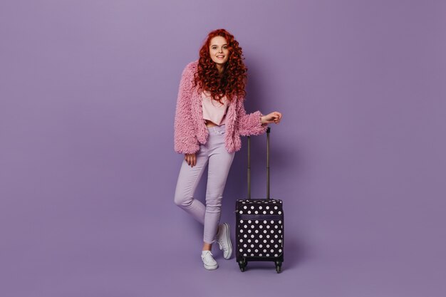 Attractive woman in light-colored pants, pink short coat and top, smiling sweetly, leaning on suitcase.