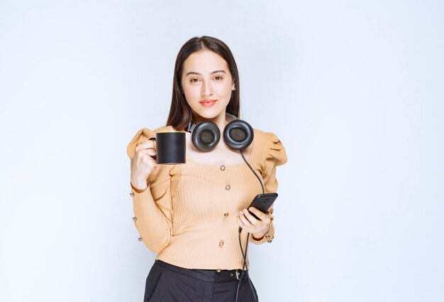 An attractive woman model holding a cup with mobile phone and headphones .